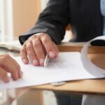 Deux personnes examinant un contrat ou un document officiel, avec une main tenant un stylo et une chemise cartonnée en arrière-plan