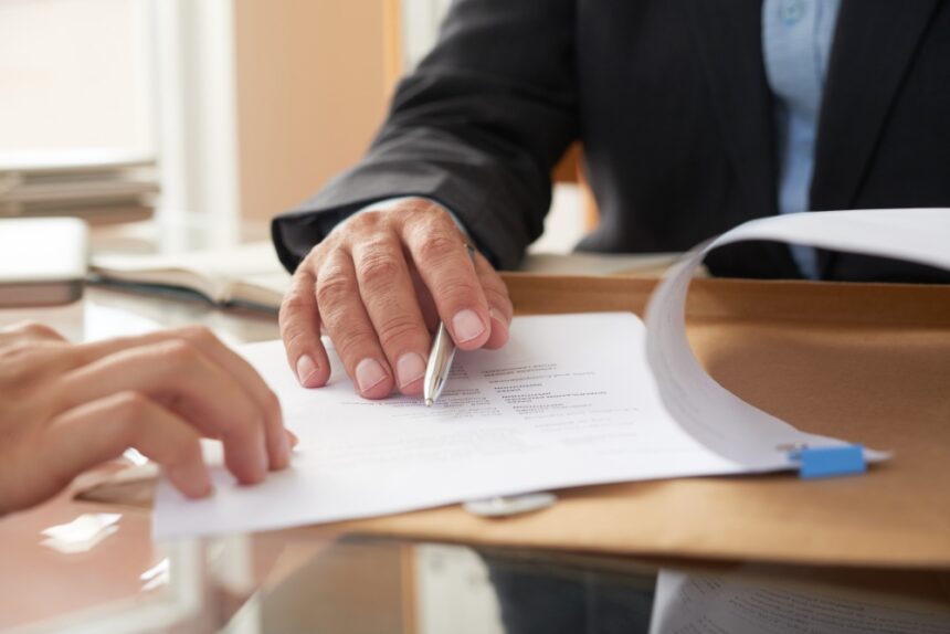 Deux personnes examinant un contrat ou un document officiel, avec une main tenant un stylo et une chemise cartonnée en arrière-plan